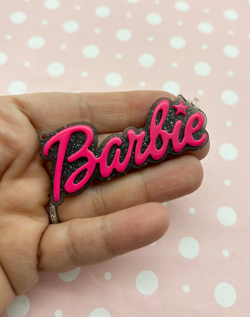 a hand holding a pink and black pin with the word barbie on it