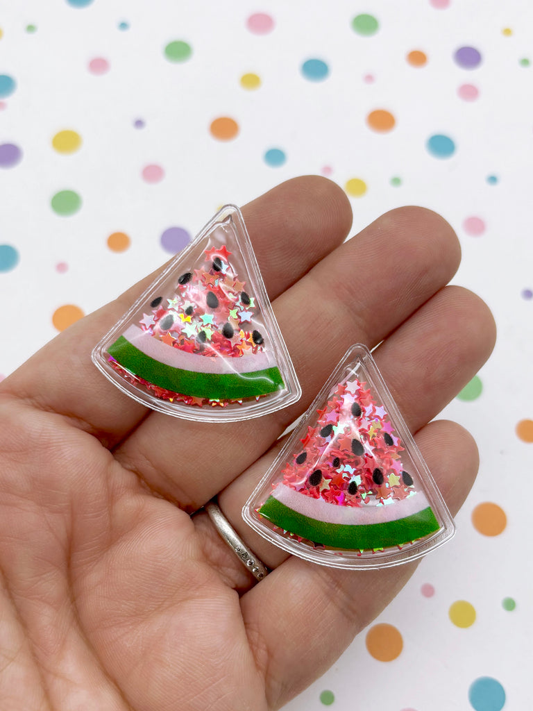 A hand holding two clear plastic earrings shaped like slices of watermelon, with glitter and black seeds on top.