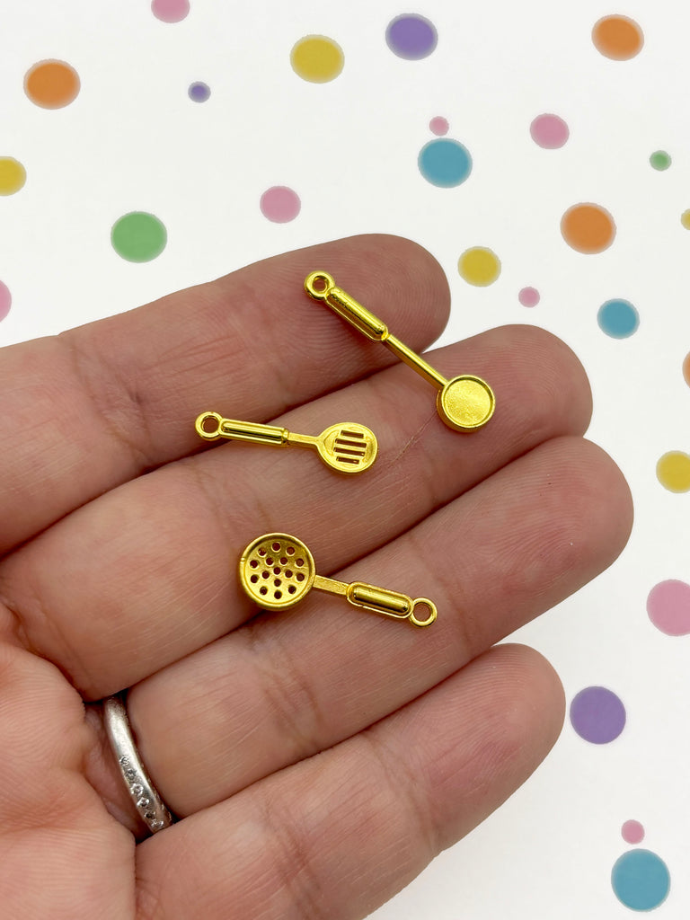 A hand holding three small gold-colored kitchen utensil charms against a background with colorful polka dots.