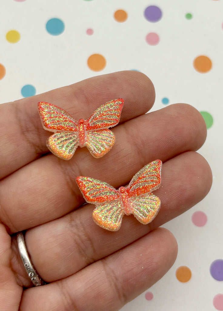 A hand holding two glittery butterfly-shaped earrings against a background with colorful polka dots.