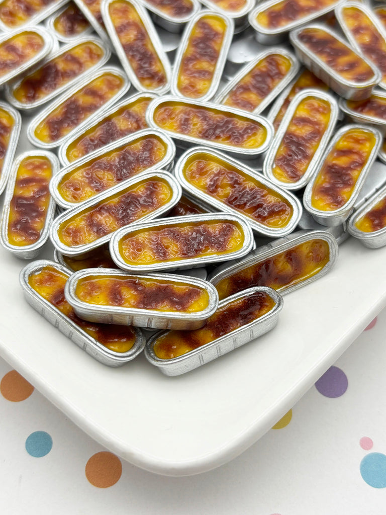 a white plate filled with small, round pastries or desserts, possibly in a tin or aluminum container. The pastries have a golden-brown color and appear to be filled with a yellow or orange substance, possibly custard or a similar filling. The plate is placed on a surface with colorful polka dots, creating a visually appealing and playful presentation.