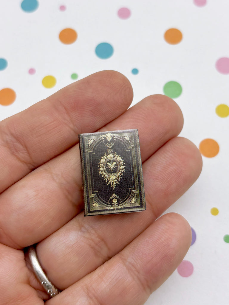 A hand holding a small, ornate book-shaped pin against a background with colorful polka dots.