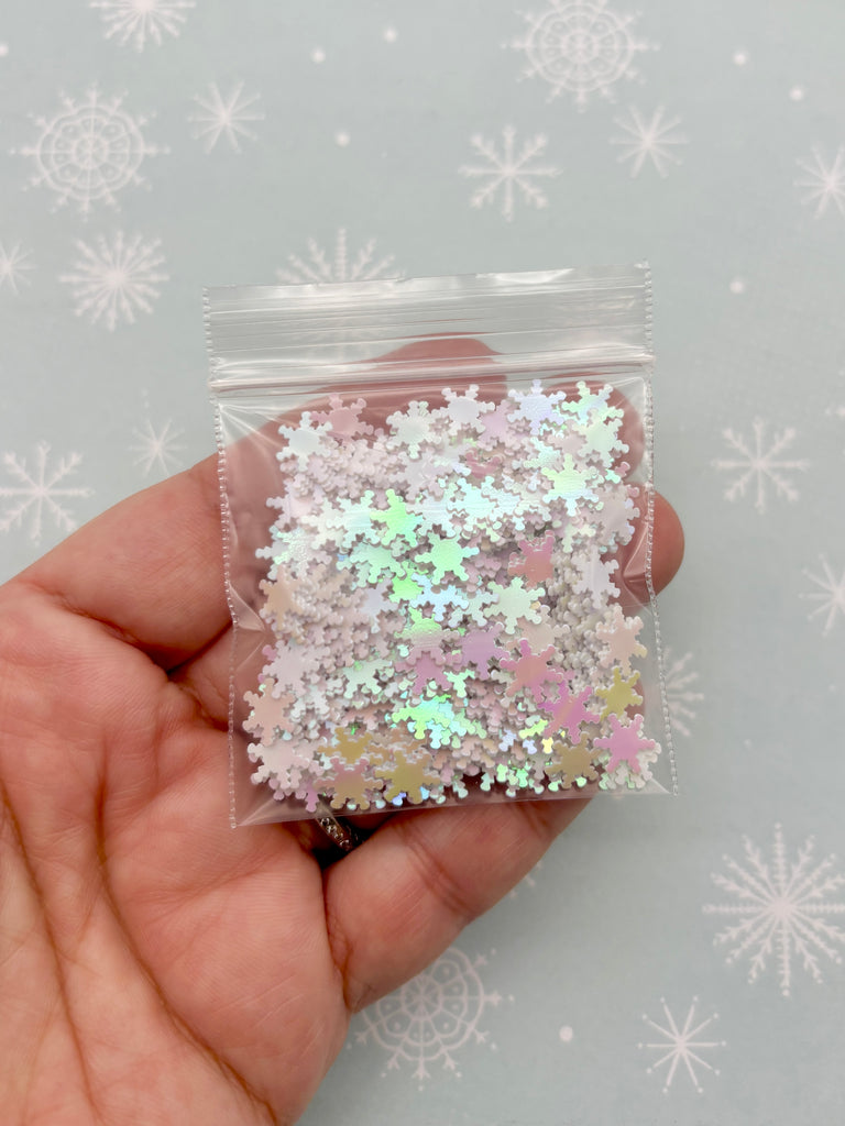 A hand is holding a clear plastic bag filled with small, multicolored glitter pieces against a background with white snowflakes.