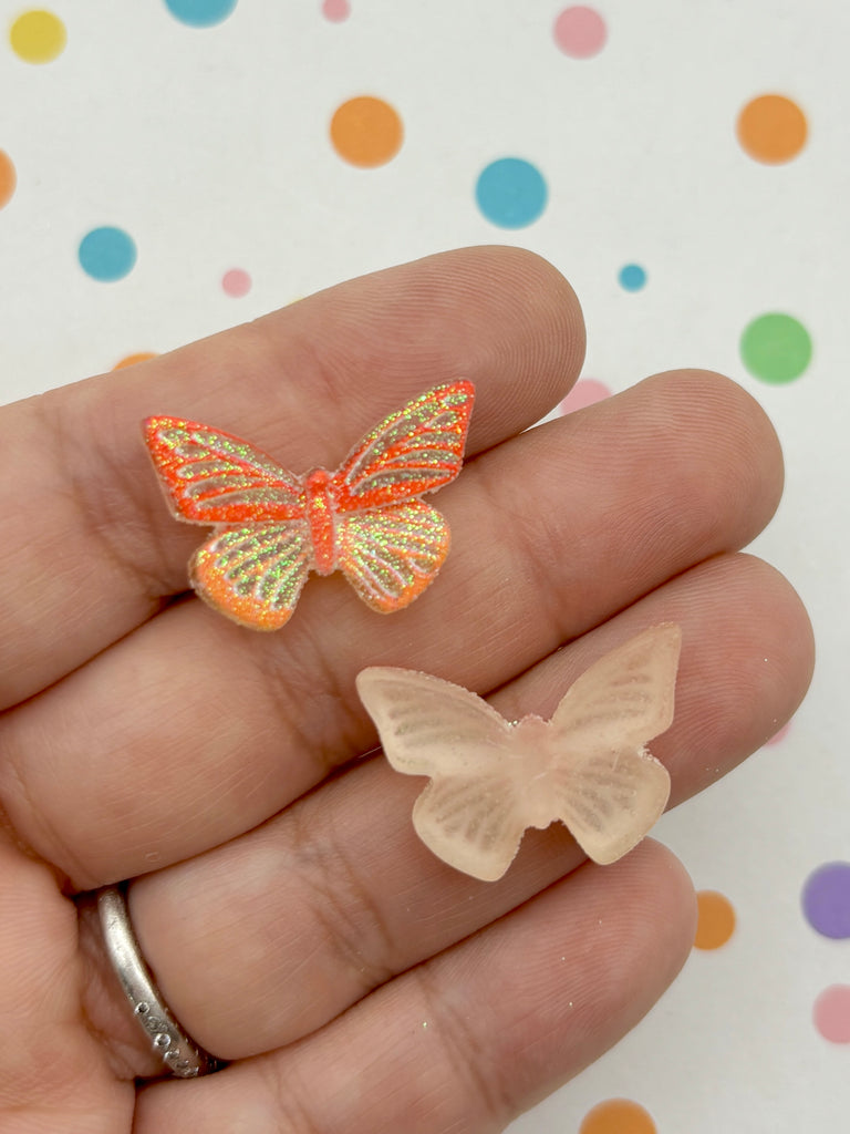 A hand holding two small butterfly-shaped objects, one with a glittery orange and yellow design and the other with a clear, light-colored design.