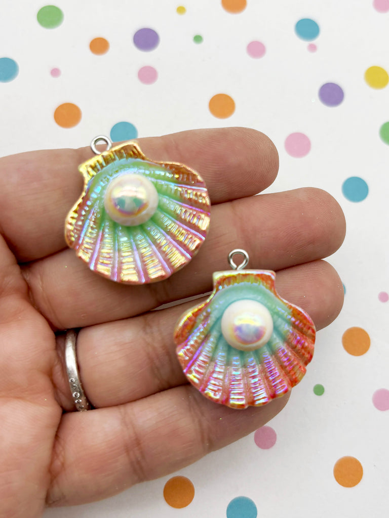 A hand holding two colorful shell-shaped pendants with a pearl in the center.