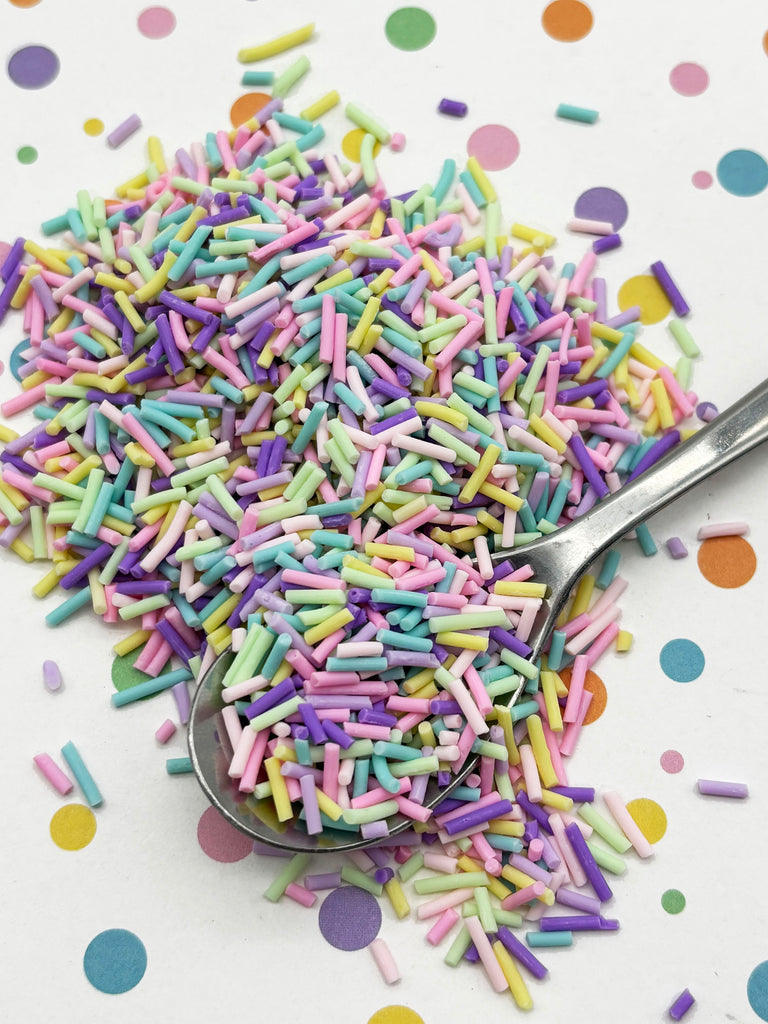 A pile of multicolored sprinkles, including pink, purple, green, and yellow, is scattered on a white surface with colorful polka dots. A spoon is visible, suggesting the sprinkles were recently used or are about to be used.
