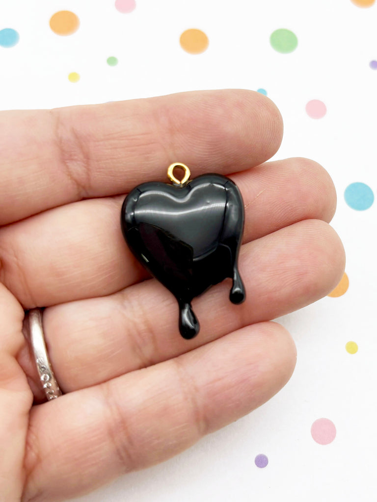 A hand holding a black heart-shaped pendant with a dripping or dripping effect, against a background with colorful polka dots.
