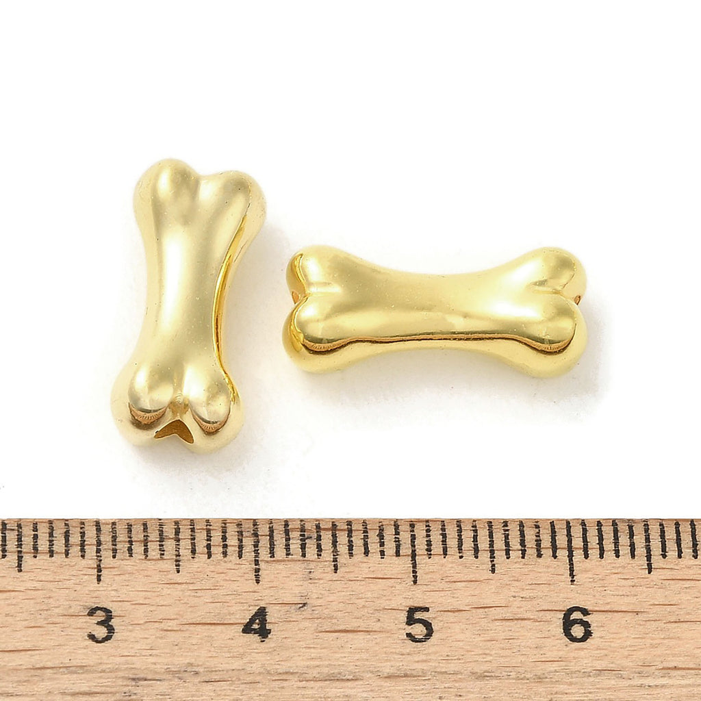 two gold-colored metal beads, which appear to be bone-shaped, placed next to a ruler for size comparison.