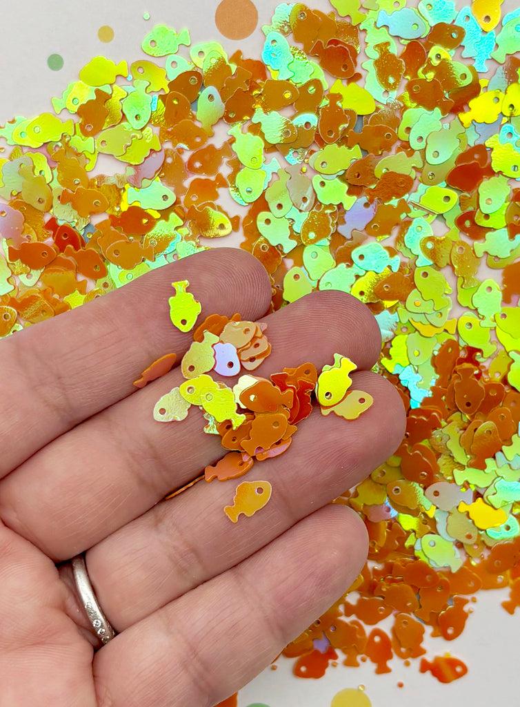 A hand holding a pile of colorful fish-shaped sequins against a background of more sequins.