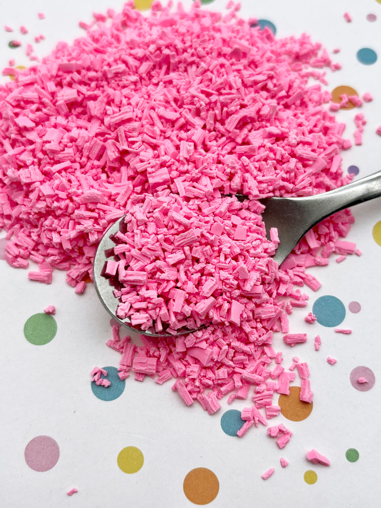 A pile of pink sprinkles on a white background, with a spoon partially submerged in the sprinkles.