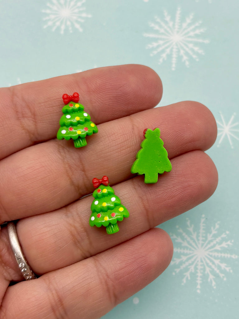 A hand holding three small green Christmas tree-shaped earrings against a light blue background with white snowflakes.