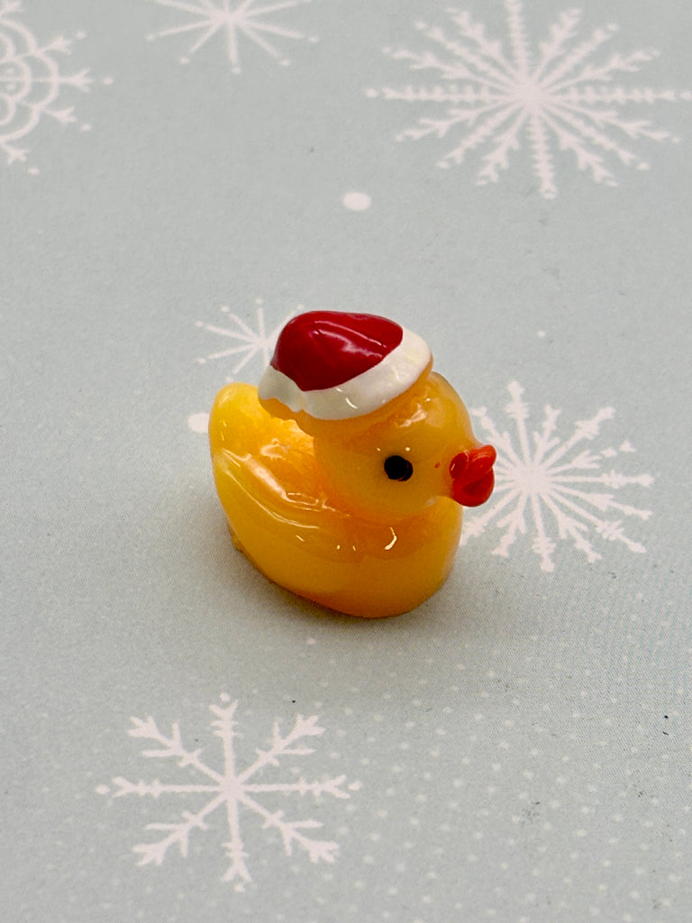 A yellow rubber duck wearing a red Santa hat is sitting on a gray background with white snowflake patterns.