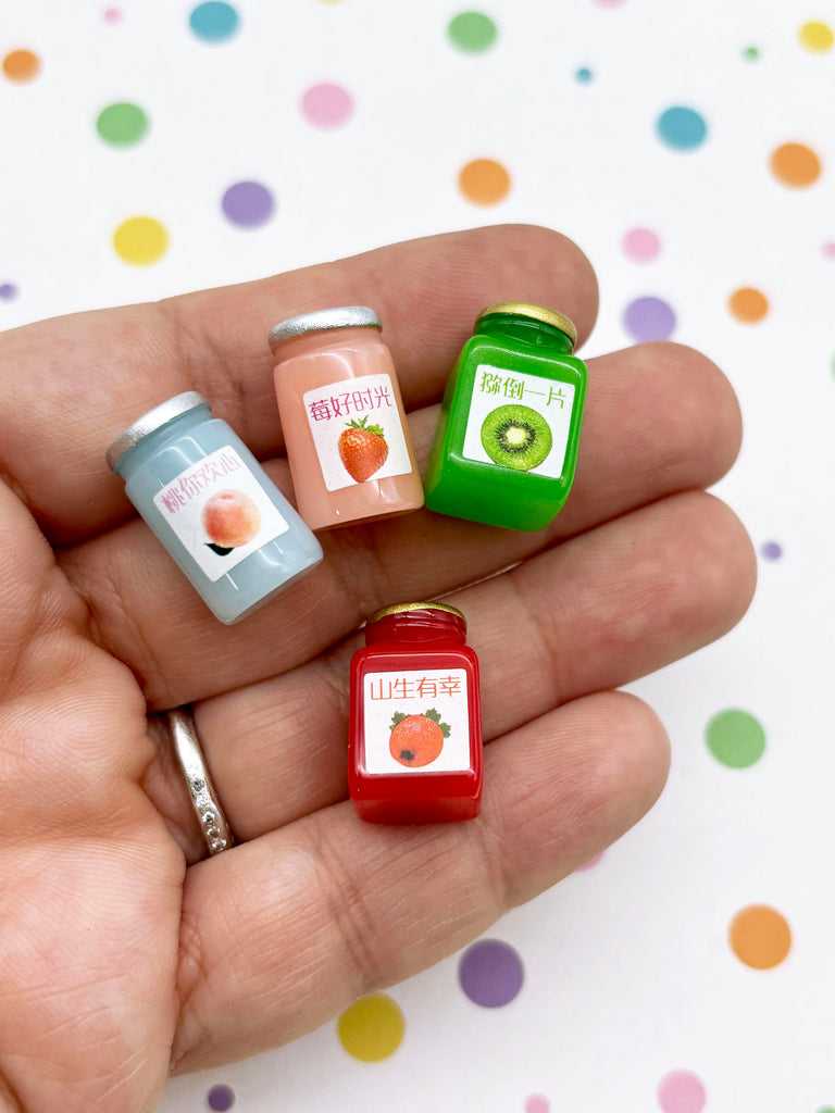 A hand holding four small jars with colorful labels, including a strawberry, kiwi, peach, and raspberry.