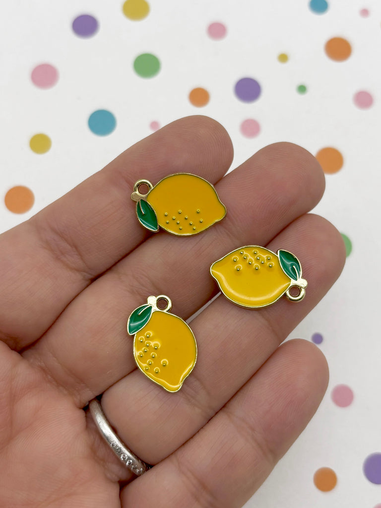 A hand holding three small, yellow enamel charms shaped like lemons against a background with colorful polka dots.