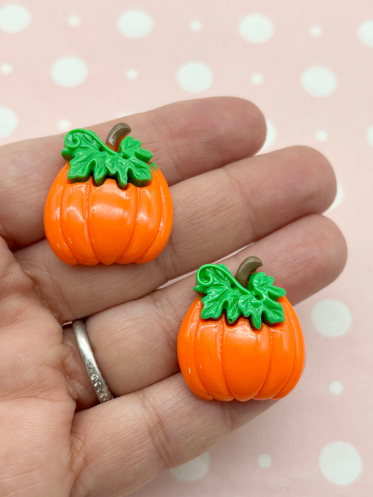 A hand holding two orange plastic pumpkins with green leaves on top against a pink background with white polka dots.