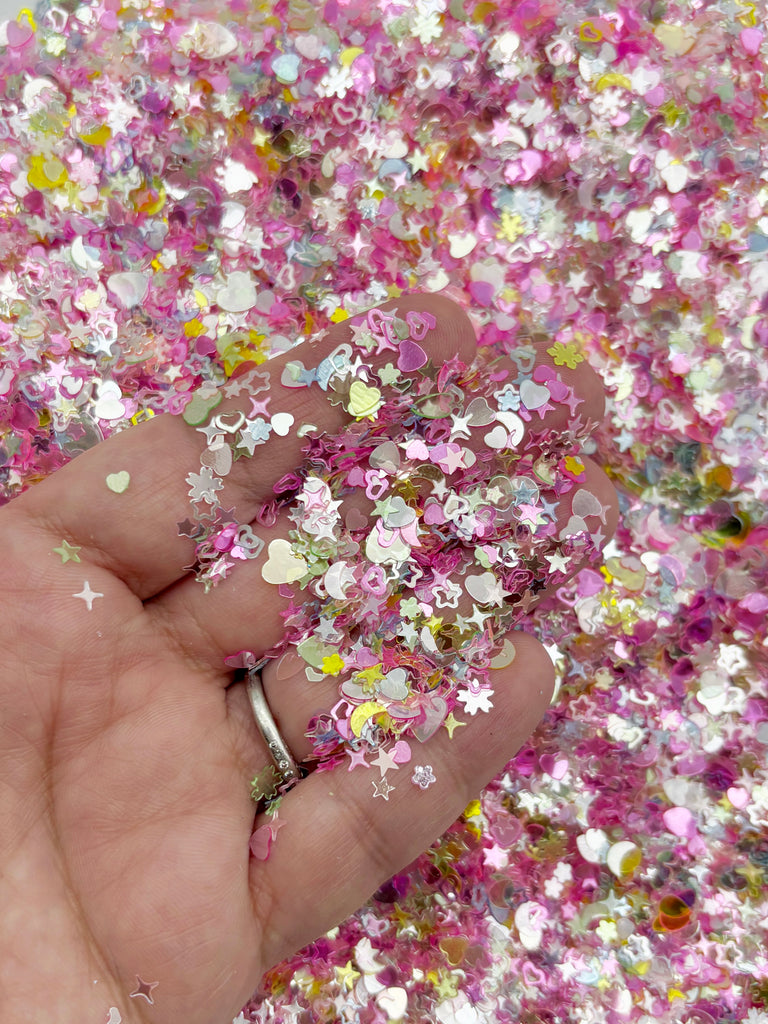 A hand is holding a pile of multicolored glitter, including pink, yellow, and white pieces, against a background of similar colors.