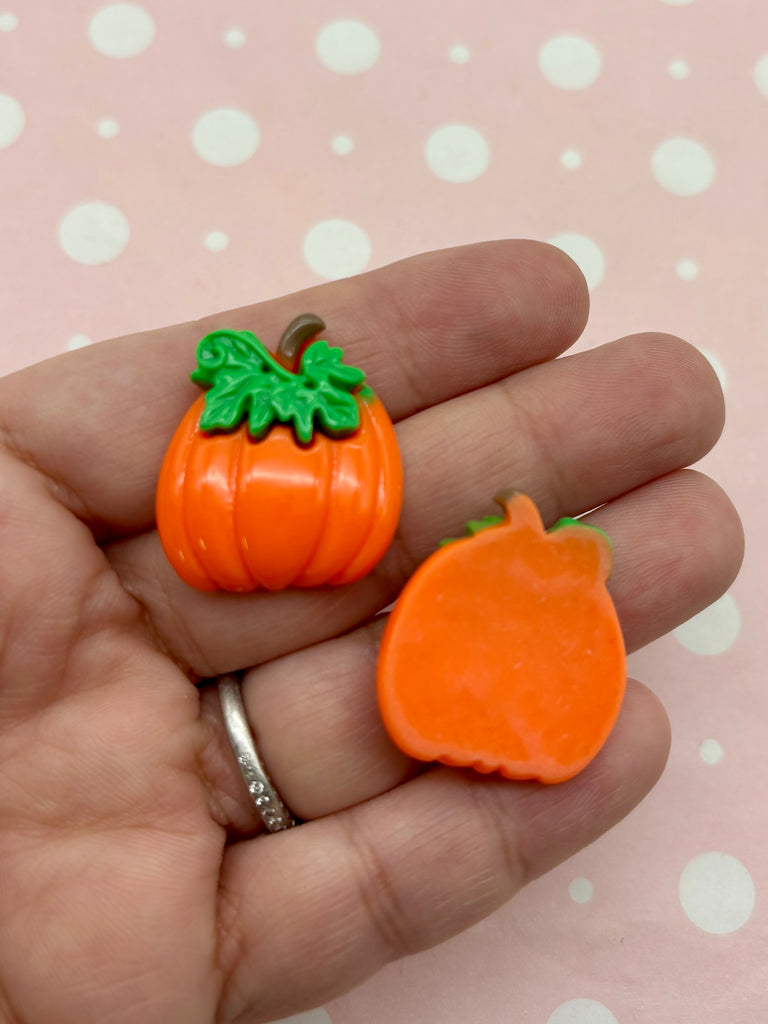 A hand holding two orange plastic pumpkins against a pink background with white polka dots.
