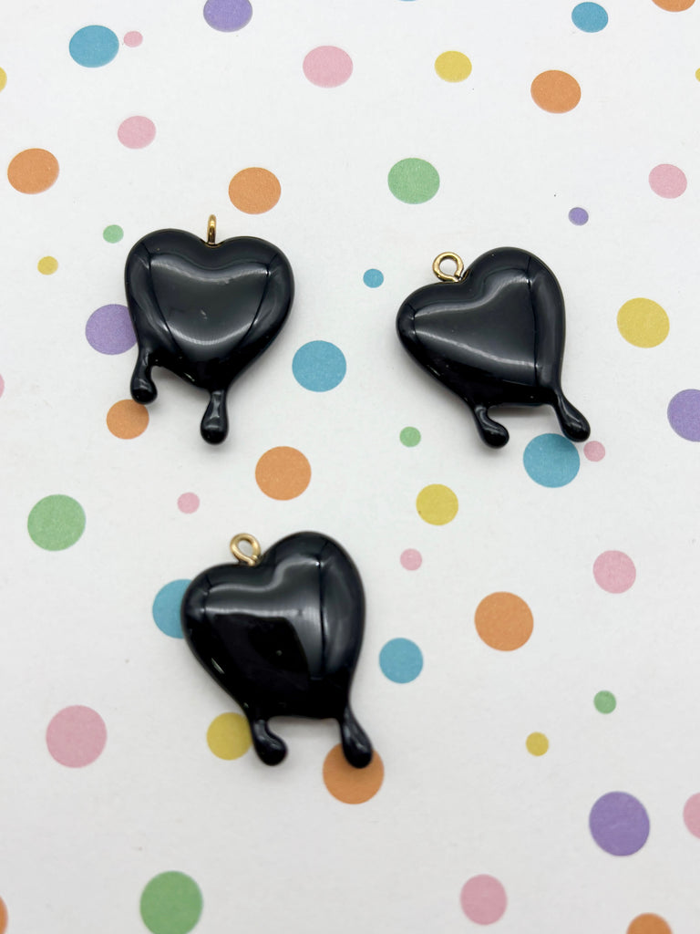 three black heart-shaped pendants with gold-colored charms, arranged on a background with colorful polka dots.