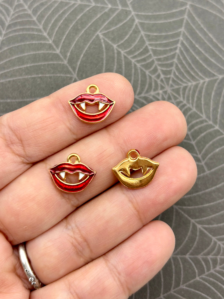 A hand holding three charms with red and gold designs, resembling teeth or lips, against a gray background.