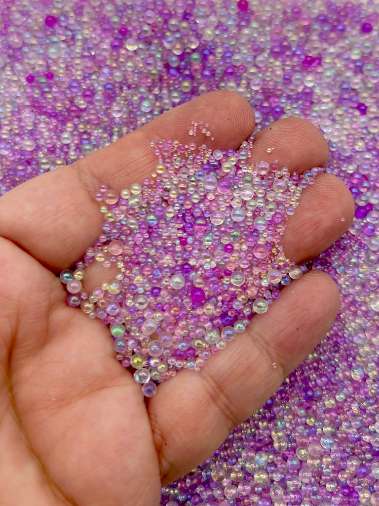 A hand is holding a pile of multicolored beads, including purple, pink, and blue, against a background of similar colored beads.
