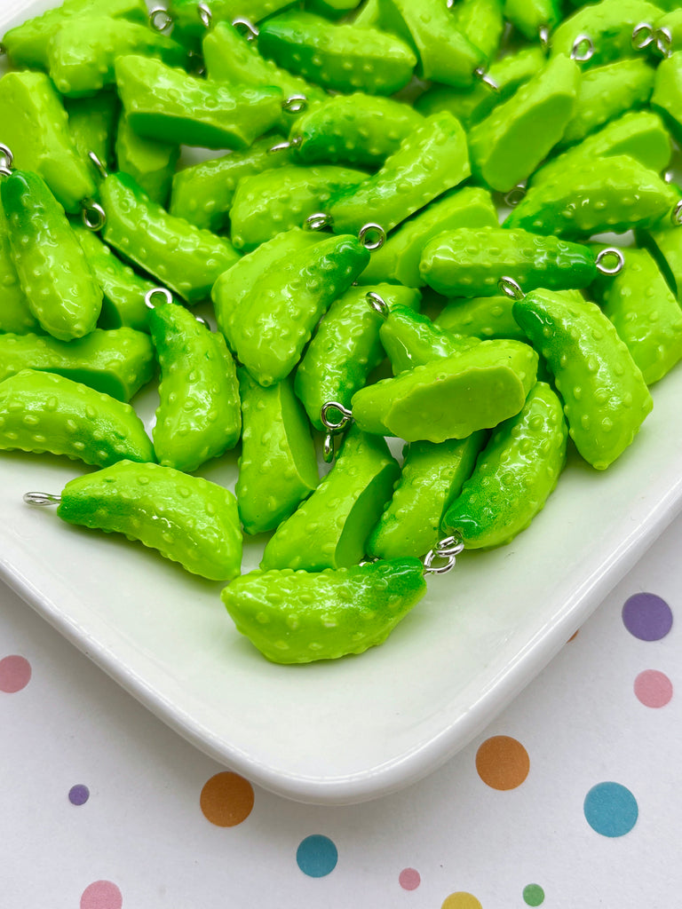 a white plate filled with bright green, plastic pickles, each with a small metal hook attached to it. The pickles are arranged in a visually appealing manner, creating a playful and colorful display.