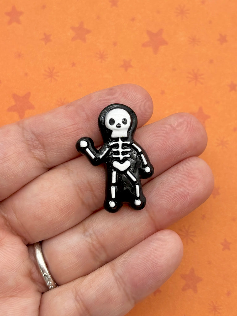 A hand holding a small black and white skeleton pin against an orange background with stars.
