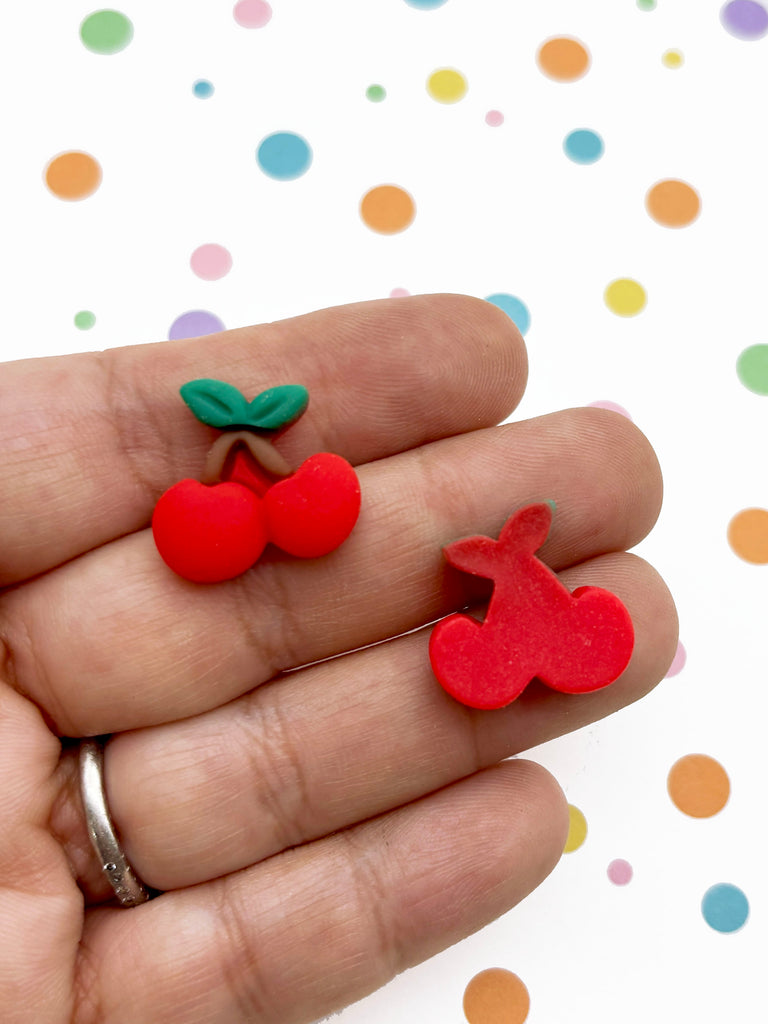 A hand holding two red plastic cherries against a background with colorful polka dots.
