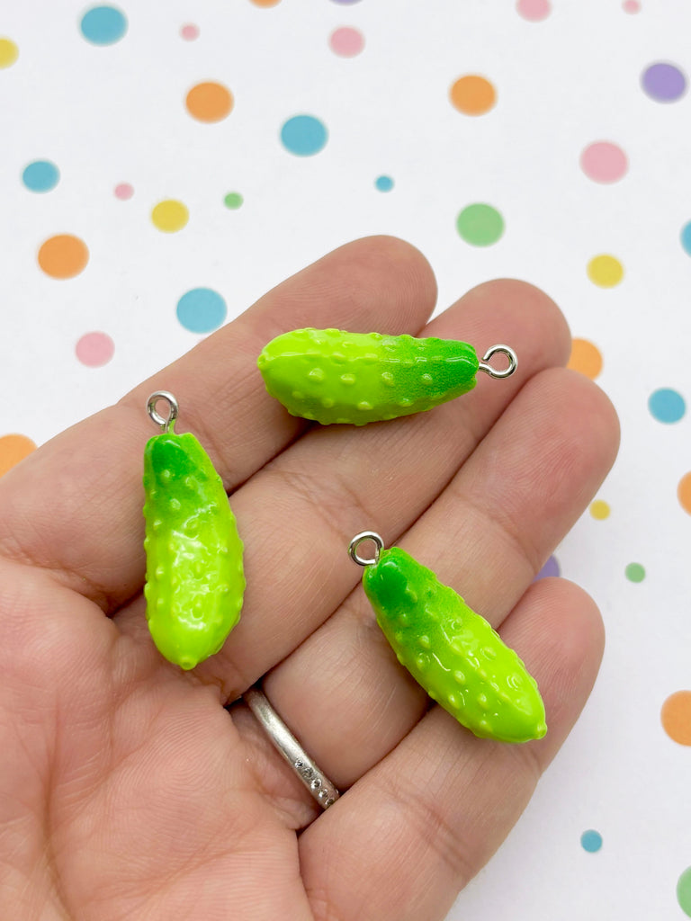 A hand holding three green, spiky cucumber-shaped charms against a background with colorful polka dots.
