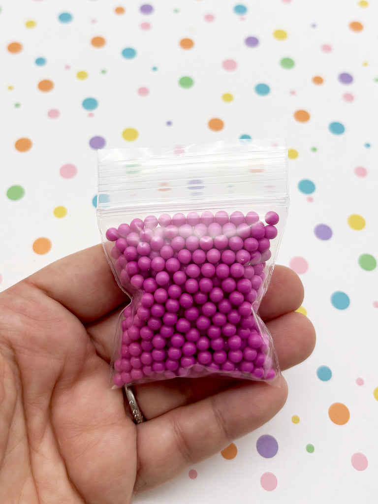 A hand is holding a small bag filled with small, round, purple beads against a background with colorful polka dots.