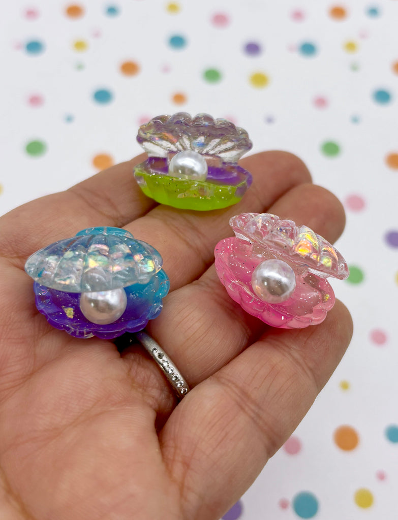 A hand holding three colorful plastic shell-shaped objects, each containing a pearl.