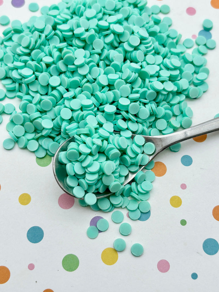 a pile of mint green confetti-like sprinkles on a white surface with colorful polka dots. A spoon is resting on the surface, and the sprinkles are spilling out of the spoon.