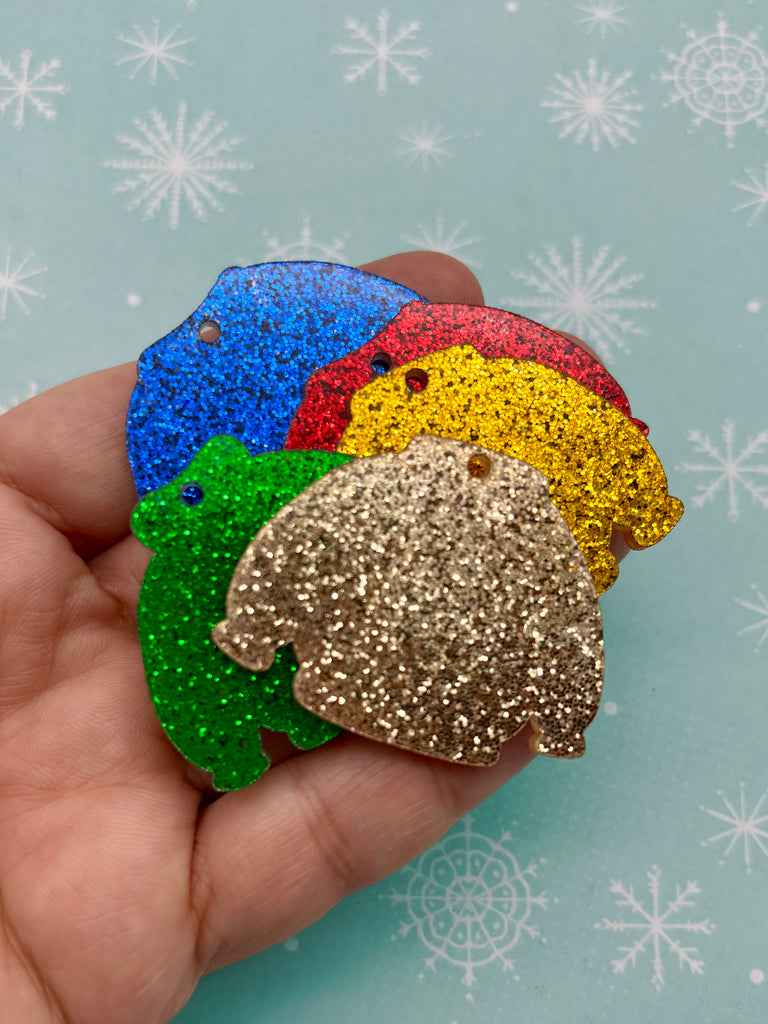 A hand is holding a collection of colorful glittery ornaments, including a green frog, a red and yellow frog, and a brown frog, against a background of snowflakes.
