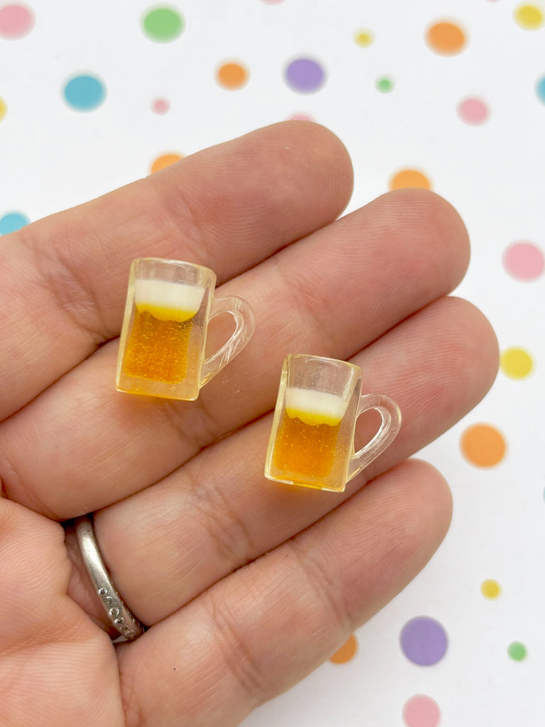 A hand holding two small, clear beer mug earrings against a background with colorful polka dots.