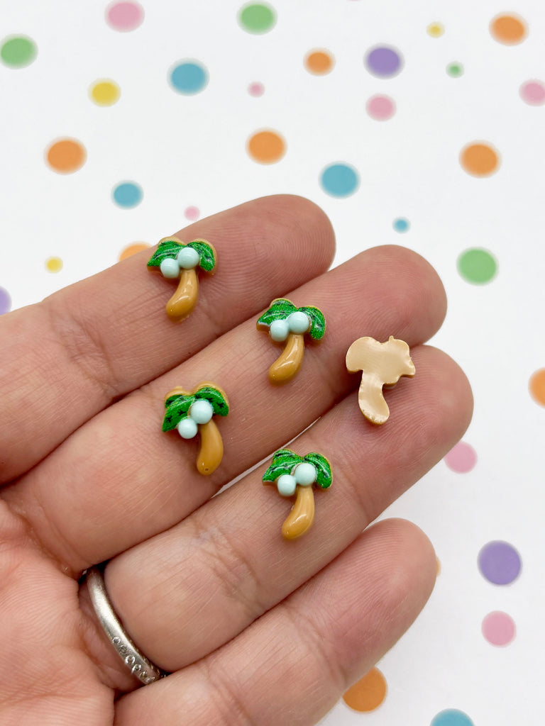 A hand holding several small, colorful earrings shaped like palm trees and bananas.