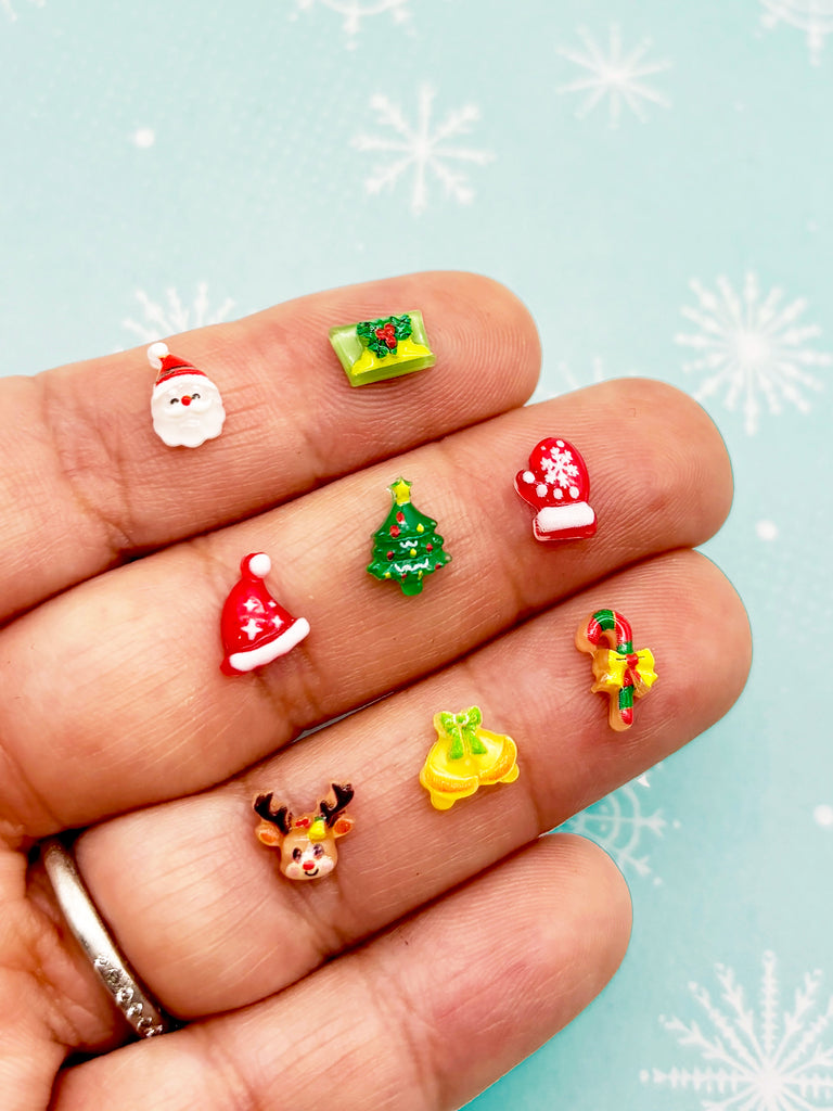 A hand holding a collection of small Christmas-themed decorations, including a Santa Claus figure, a Christmas tree, a red Santa hat, a green Christmas tree, a red Santa hat, a yellow bell, a reindeer, and a candy cane.