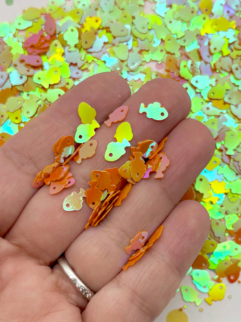 A hand holding a collection of small, colorful fish-shaped sequins against a background of similar sequins.