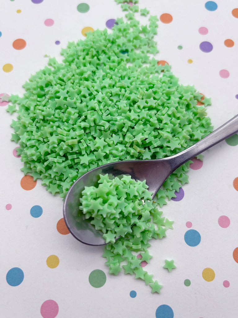 a pile of green star-shaped sprinkles on a white surface with colorful polka dots. A spoon is resting on top of the sprinkles, and the sprinkles are scattered around the spoon.