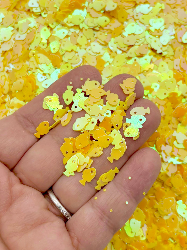 A hand holding a pile of yellow glittery objects, resembling small fish or sea creatures, against a background of similar yellow glittery objects.