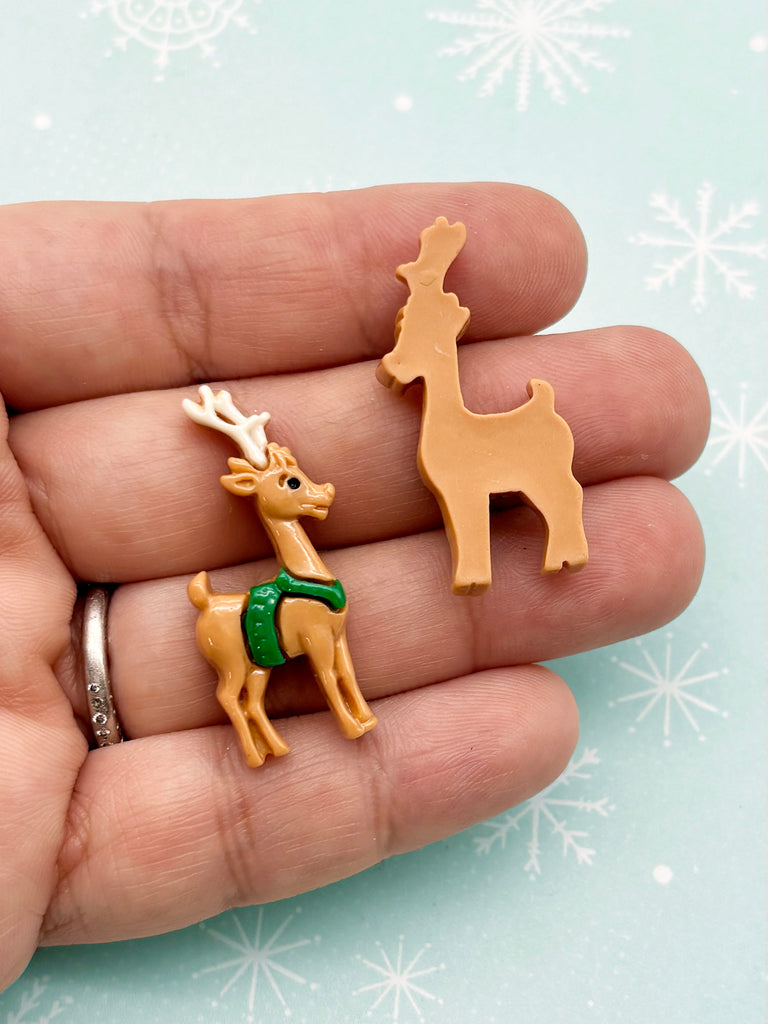 A hand holding two small toy reindeer figures against a snowy background.