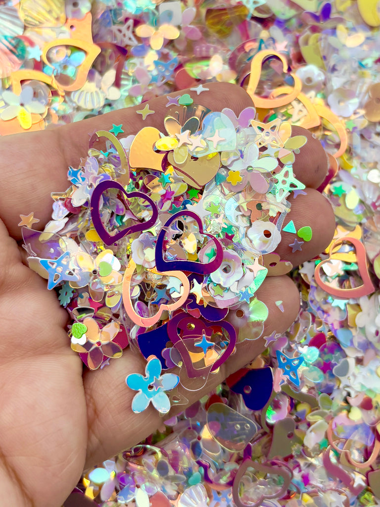 A hand is holding a pile of colorful, glittery, and shiny objects, including hearts, flowers, and stars, which appear to be confetti or small decorative pieces.