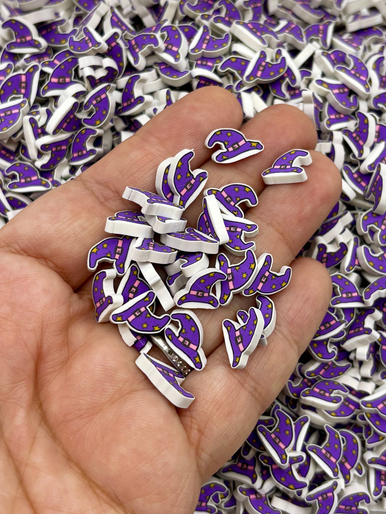 A hand holding a pile of small purple and white beads, which appear to be witch's hats or similar accessories.