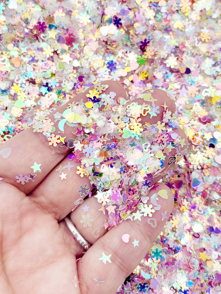A hand is holding a pile of multicolored glitter, including various shapes like stars, hearts, and flowers, creating a vibrant and festive atmosphere.