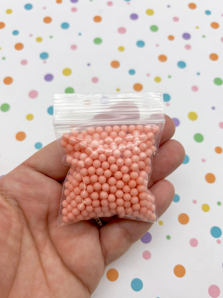 A hand holding a small bag filled with small, round, pink beads against a background with colorful polka dots.