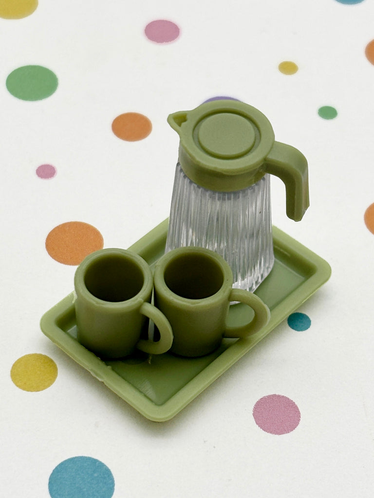 A green plastic pitcher and two matching green cups are arranged on a green tray, placed on a white surface with colorful polka dots.