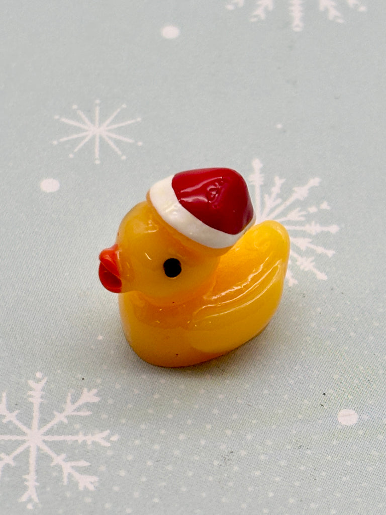 A yellow rubber duck wearing a red Santa hat is sitting on a light blue background with white snowflake patterns.