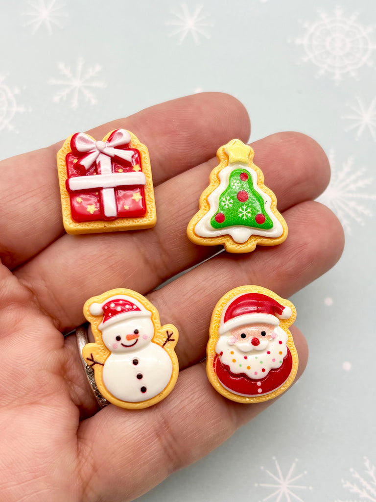 A hand holding four small Christmas-themed cookies or candies, including a gift box, a Christmas tree, a snowman, and a Santa Claus cookie.