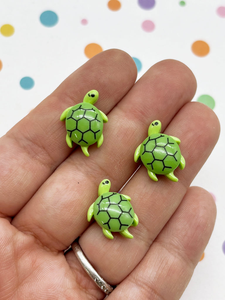 A hand holding three small green turtle-shaped charms against a background with colorful polka dots.