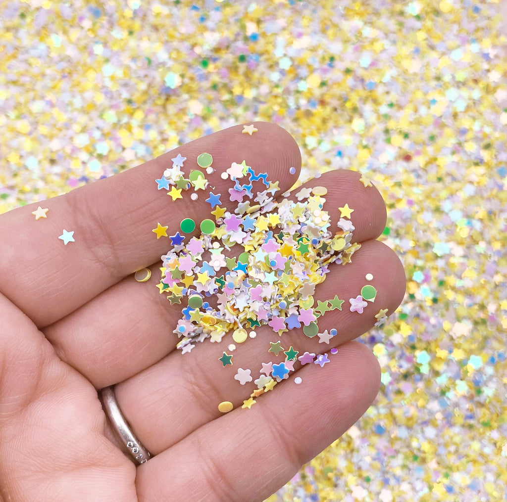 A hand holding a pile of colorful star-shaped glitter pieces against a background of yellow and pink glitter.