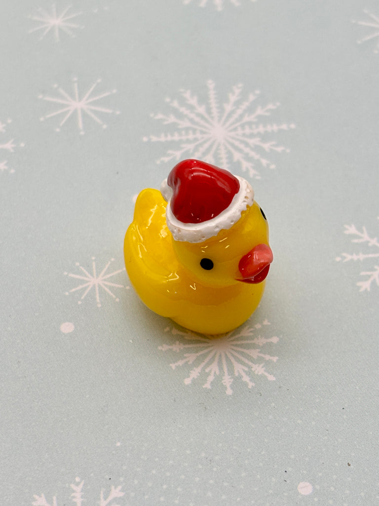 A yellow rubber duck wearing a red Santa hat is sitting on a light blue background with white snowflake patterns.