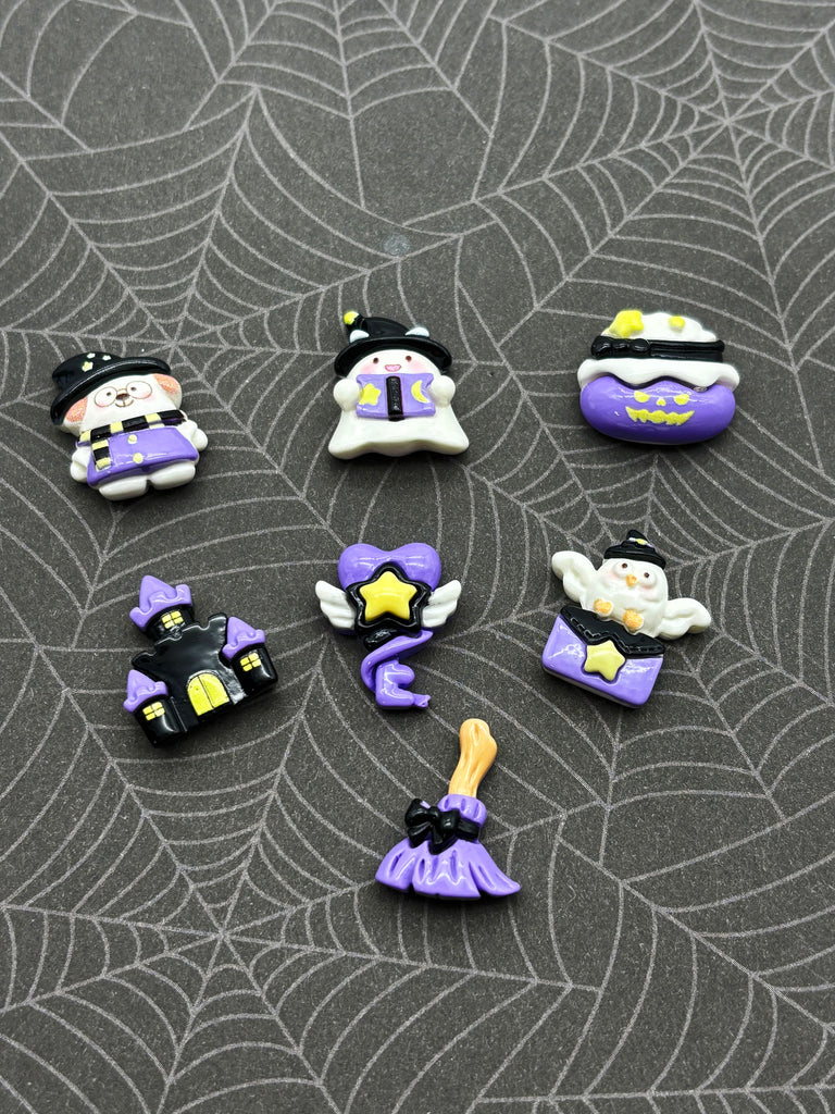 a collection of Halloween-themed small figurines and decorations, including a witch, ghost, pumpkin, spider, and broomstick, arranged on a spider web patterned background.